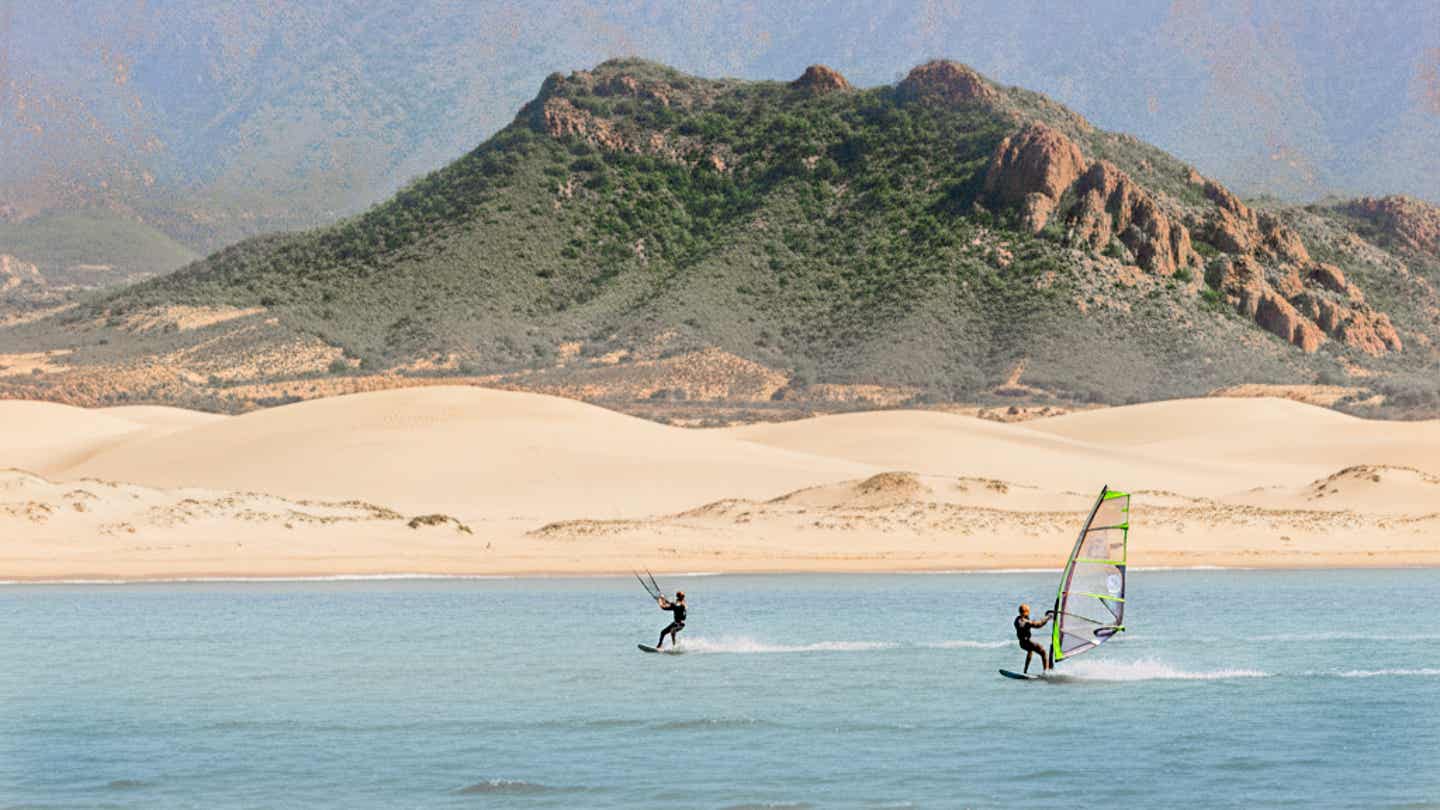 Windsurfer und Kitesurfer gleiten vor den Dünen von Tarifa über das Wasser mit Blick auf grüne Berglandschaft 