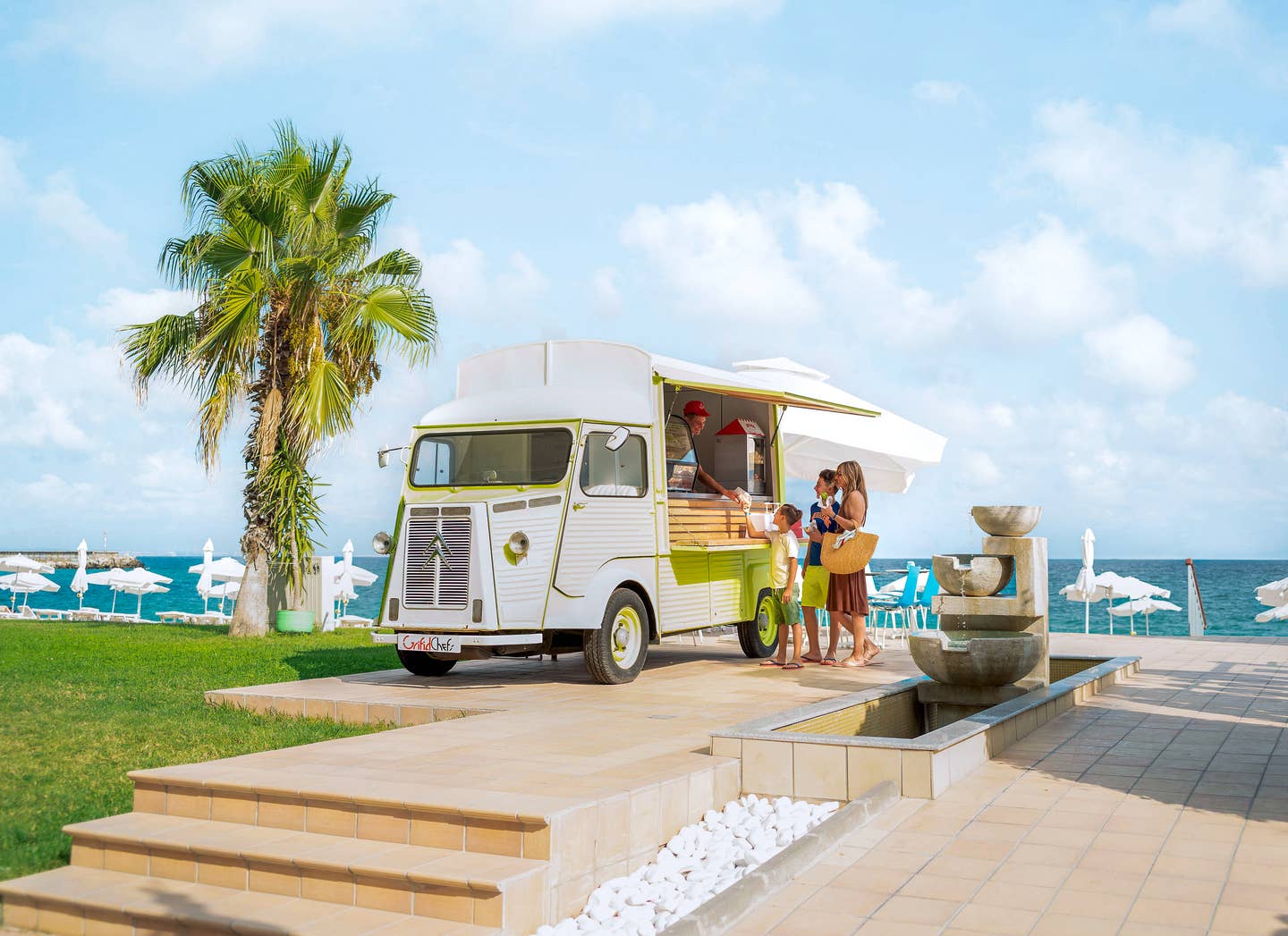 Foodtruck mit offener Theke am Strand neben einer Palme und einer kleinen Wasserfontäne, davor stehen drei Personen