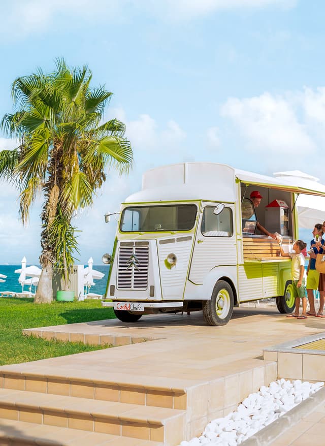 Sentido Familienhotels Foodtruck mit offener Theke am Strand neben einer Palme und einer kleinen Wasserfontäne, davor stehen drei Personen