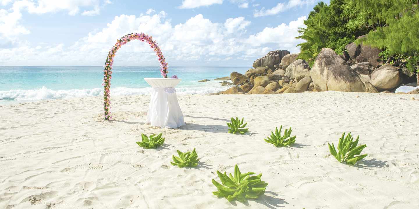 Blumenbogen und mit weißem Tuch bedeckter Tisch auf einem Sandstrand mit grünen Pflanzen und Felsen im Hintergrund