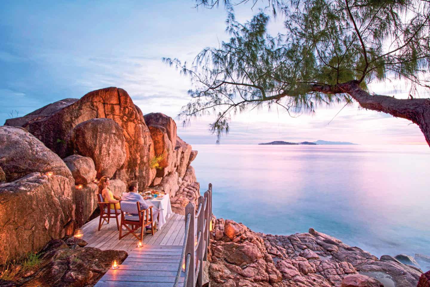 Ein Paar macht Urlaub im Indischen Ozean, sitzt auf einem Holzsteg zwischen Felsen mit Blick auf das Meer und genießt den Sonnenuntergang auf den Seychellen bei einem exklusiven Essen