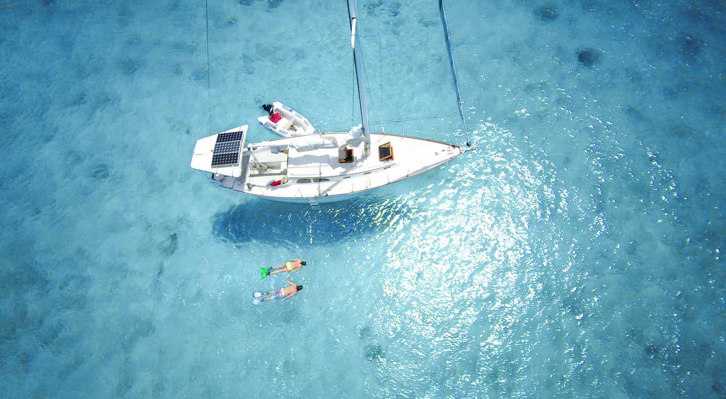 Paar schnorchelt Hand in Hand neben einem Segelboot mit Solarpanel in türkisfarbenem Wasser der Karibik aus der Luft