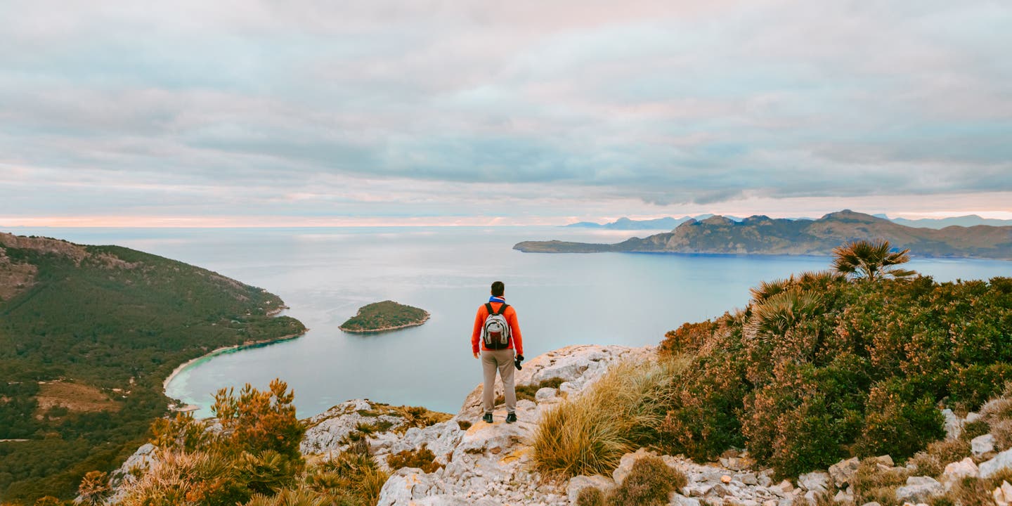 Junger Wanderer schaut morgens mit Fernglas vor der Nordküste Mallorcas in die Ferne - Bitte Al