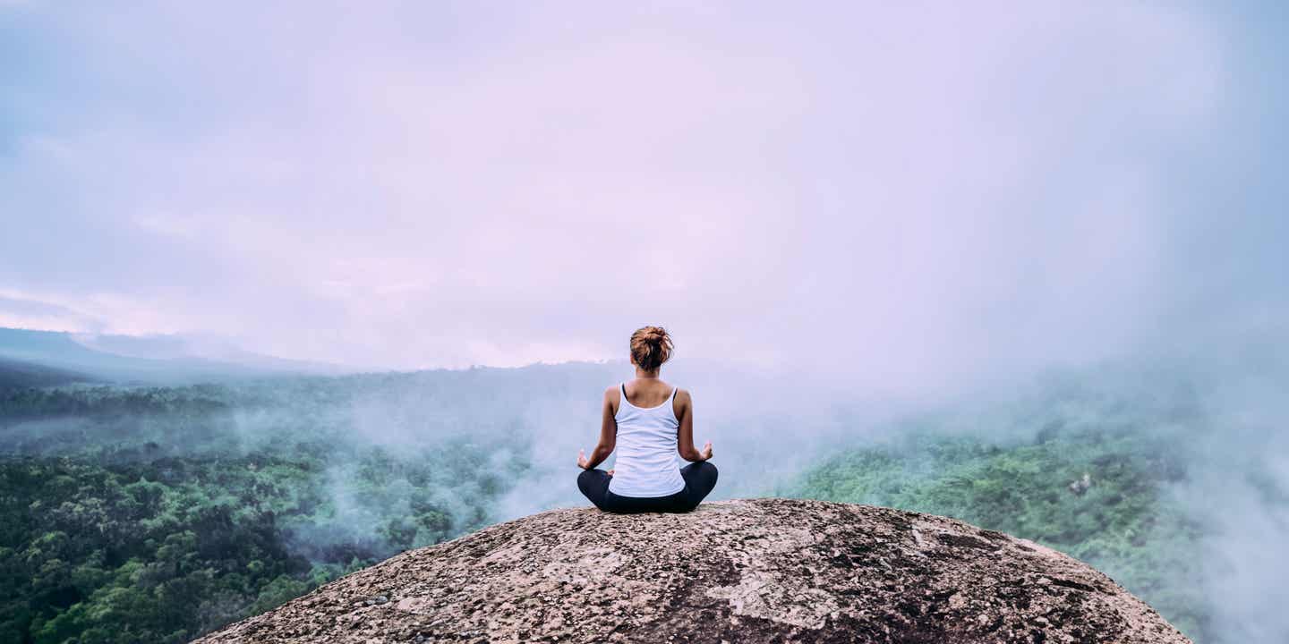 Frau sitzt in Yoga-Pose auf einem Felsen und blickt über den Dschungel Thailands
