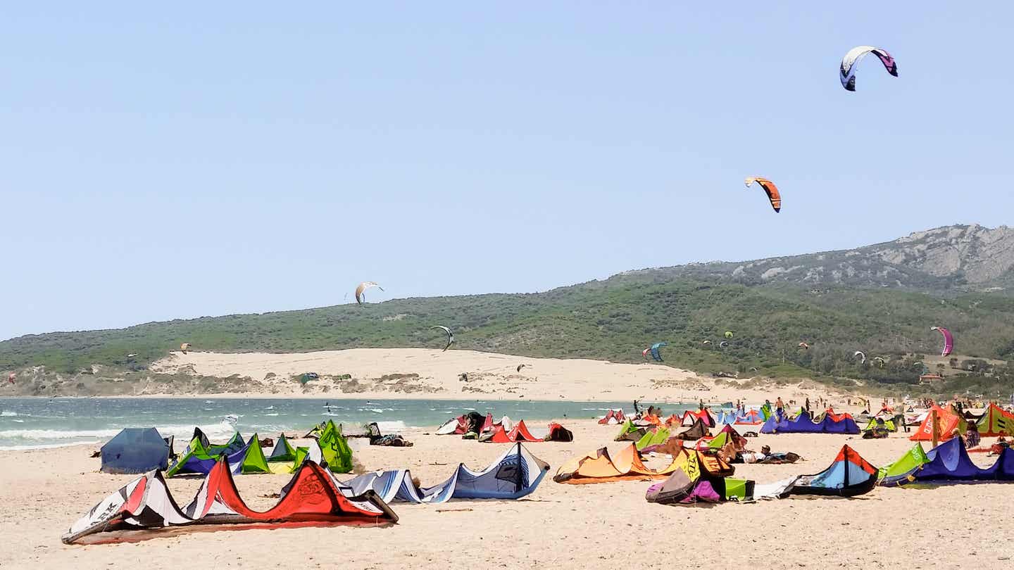 Kites liegen am Strand von Tarifa, während bunte Schirme über dem Meer in der Luft tanzen 