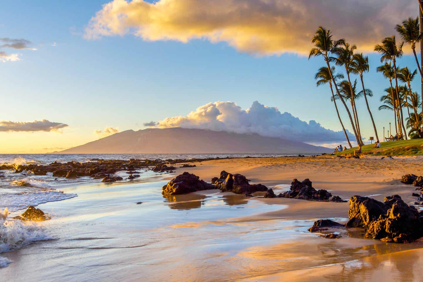 Sonnenuntergang an einem Strand auf Maui mit sanften Wellen, Felsen, Palmen und Bergen.