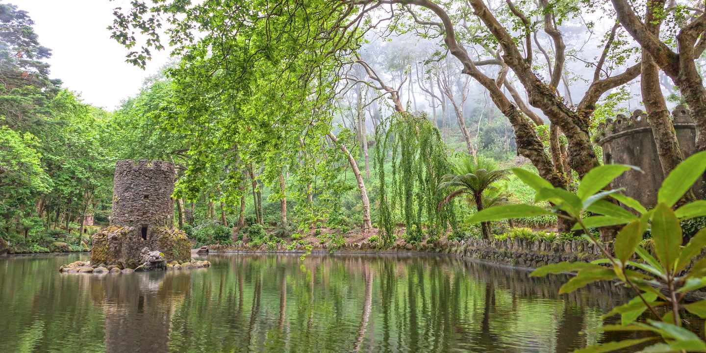 Parkanlage in Sintra mit Teich, kleinem Steinturm und dichtem Baumbestand im Palacio Nacional de Pena