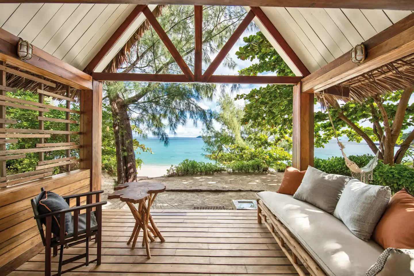 Veranda im Constance Tsarabanjina mit Blick auf tropische Pflanzen und weißen Sandstrand Offene Veranda eines Bungalows im Constance Tsarabanjina, Madagaskar, mit Sofa, Hängematte und Blick auf den Indischen Ozean