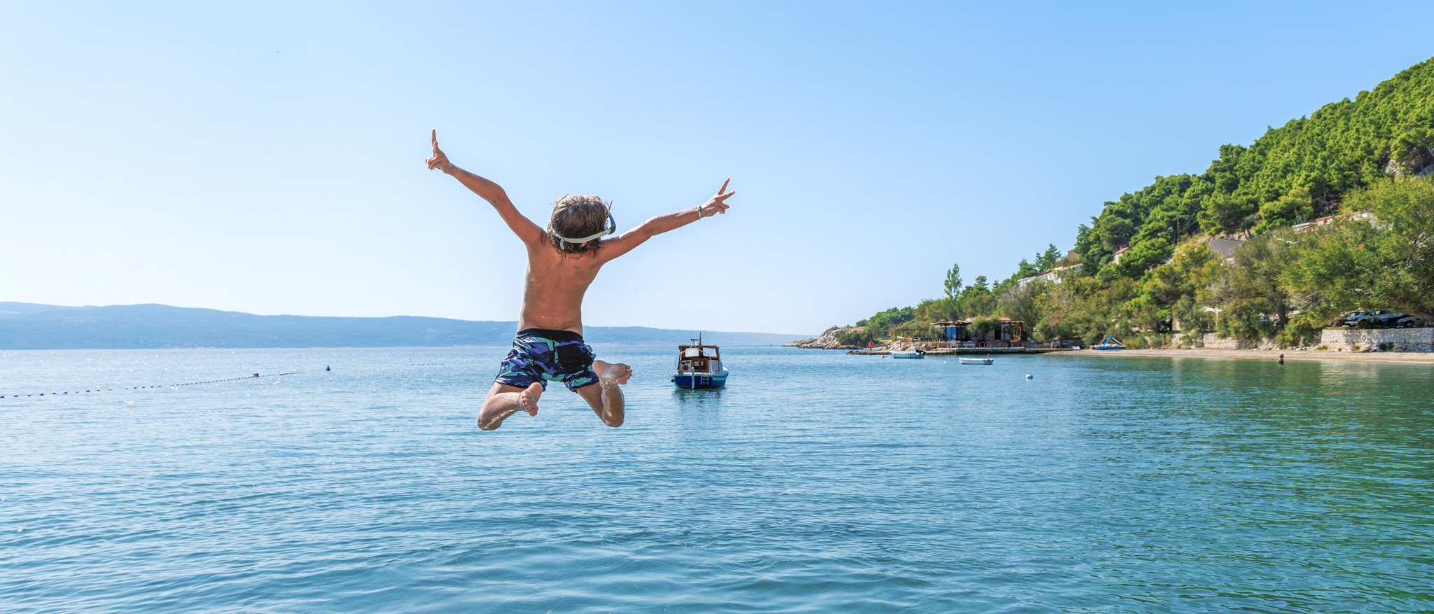 Reiseziele mit Kindern: Junge springt an einem sonnigen Urlaubstag von einem Steg ins türkisblaue Meer von Kroatien