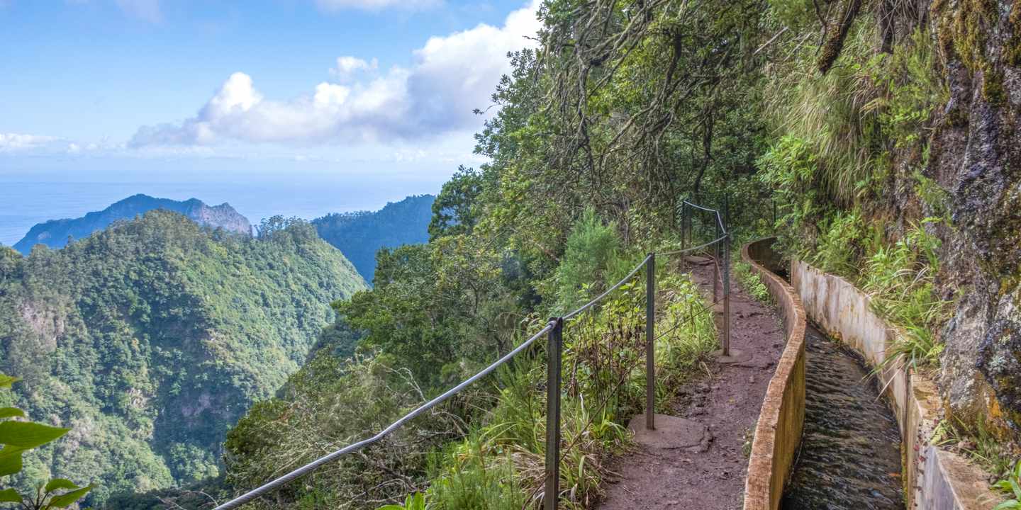 Schmaler Wanderweg entlang der Levada do Furado mit Geländer und Blick über grüne Berglandschaft