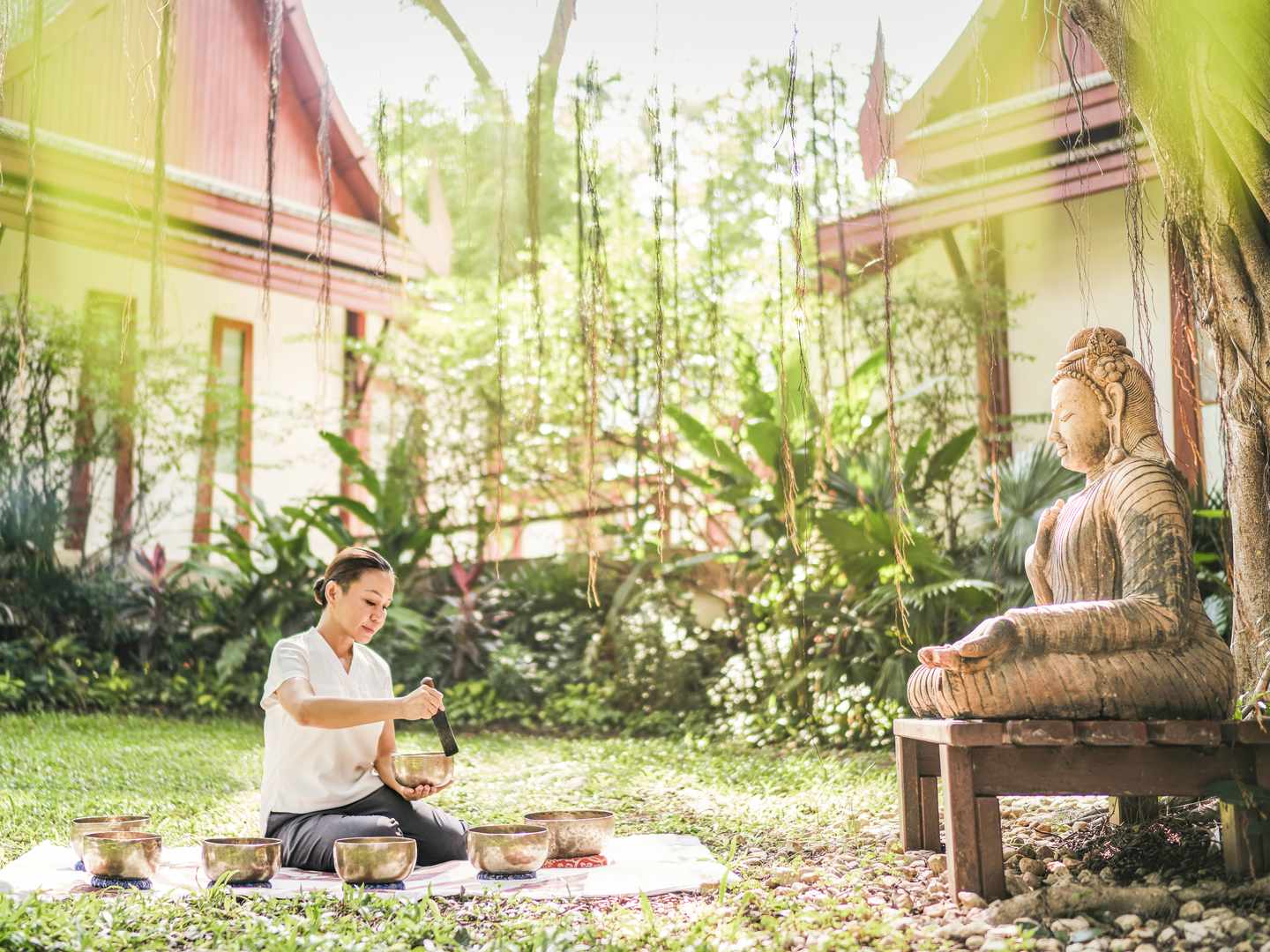 Thailändische Wellness-Rituale Frau mit Klangschalen sitzt im Grad vor einer steinernen Buddha-Figur