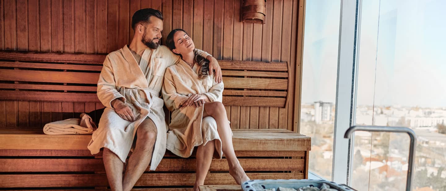Wellnessurlaub mit einem Paar im Bademantel, das gemeinsam in einer Sauna mit Panoramafenster entspannt Wellnessurlaub mit einem Paar im Bademantel, das gemeinsam in einer Sauna mit Panoramafenster entspannt