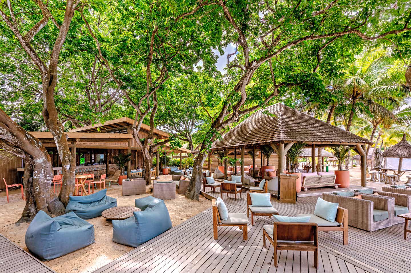 Blick auf eine Sitzlandschaft im The Ravenala Attitude auf Mauritius mit zahlreichen Holzmöbeln unter imposanten Bäumen
