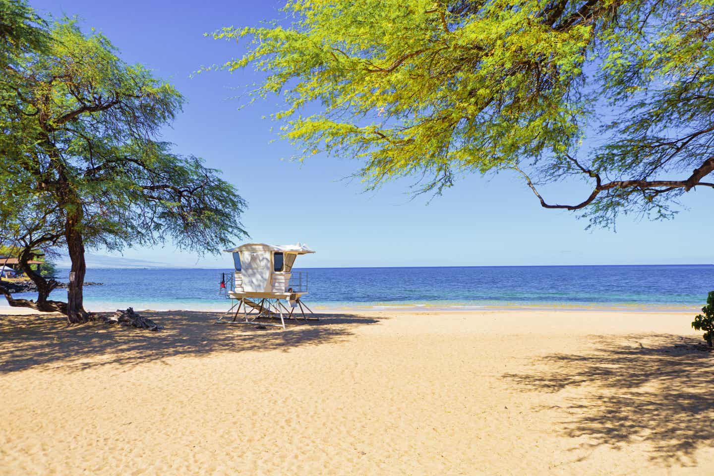 Blick auf Hawaiis Strand Hapuna Beach mit weißem Rettungsschwimmerturm im feinen Sand und grünen Bäumen vor blauem Meer und Himmel.