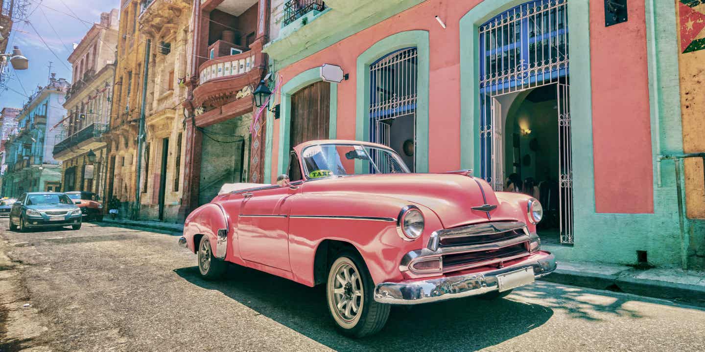 Ein knallpinkes Oldtimer-Taxi steht in einer Straße mit bunten Häuserfassaden in Havanna auf Kuba