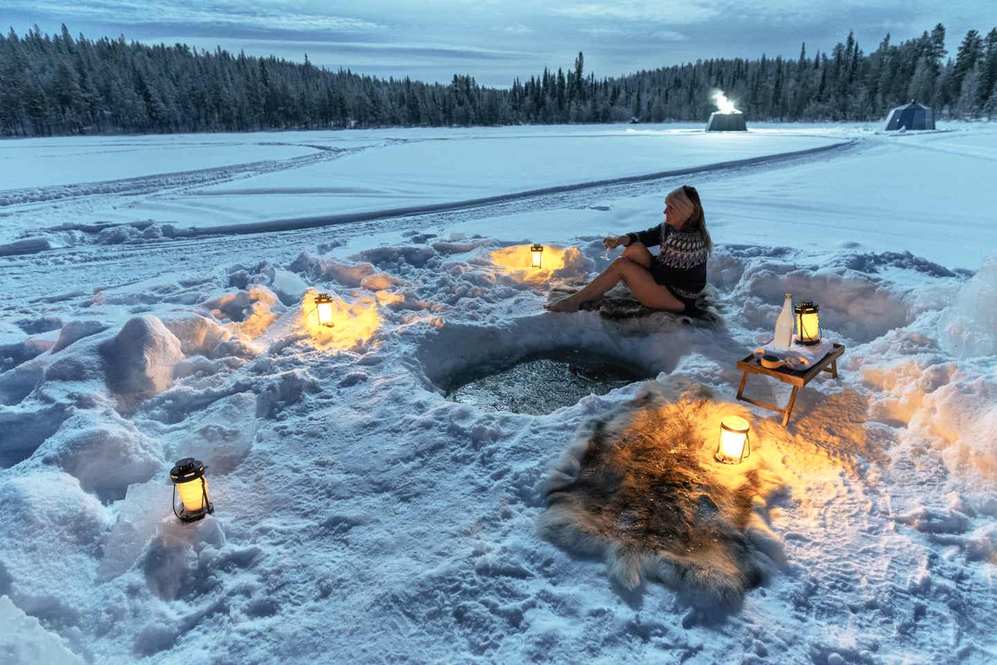 Schweden Urlaub mit DERTOUR. Frau sitzt mit Drink und Laternen abends an einem Loch zum Eisbaden