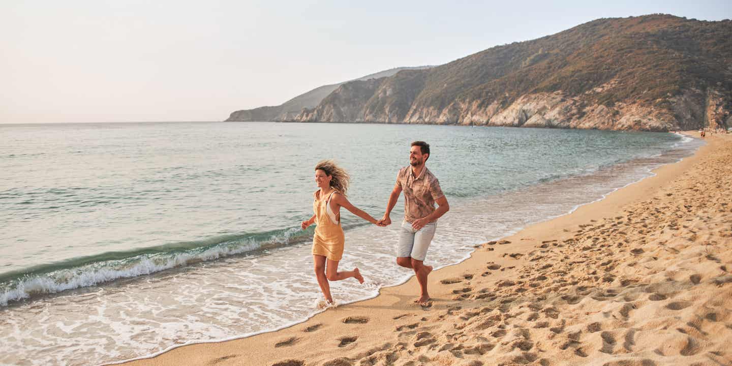 So geht Strandurlaub in Griechenland: ein Paar geht an einem malerischen Strand spazeiren