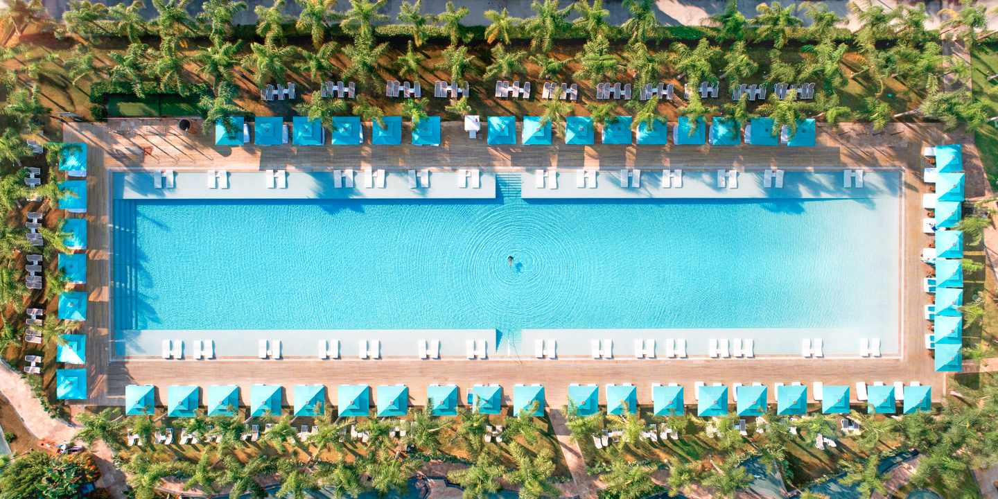 Vogelperspektive auf länglichen Pool umrandet mit Sonnenschirmen, Liegen und Palmen