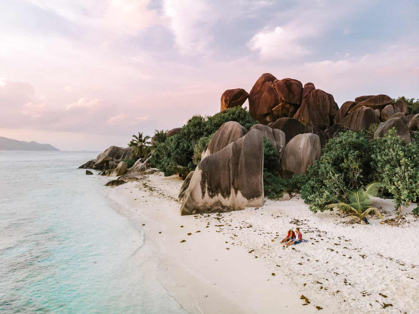 Ein Paar sitzt im Sand am Strand Anse Source d’Argent im La Digue Urlaub, im Hintergrund Palmen und Granitfelsen