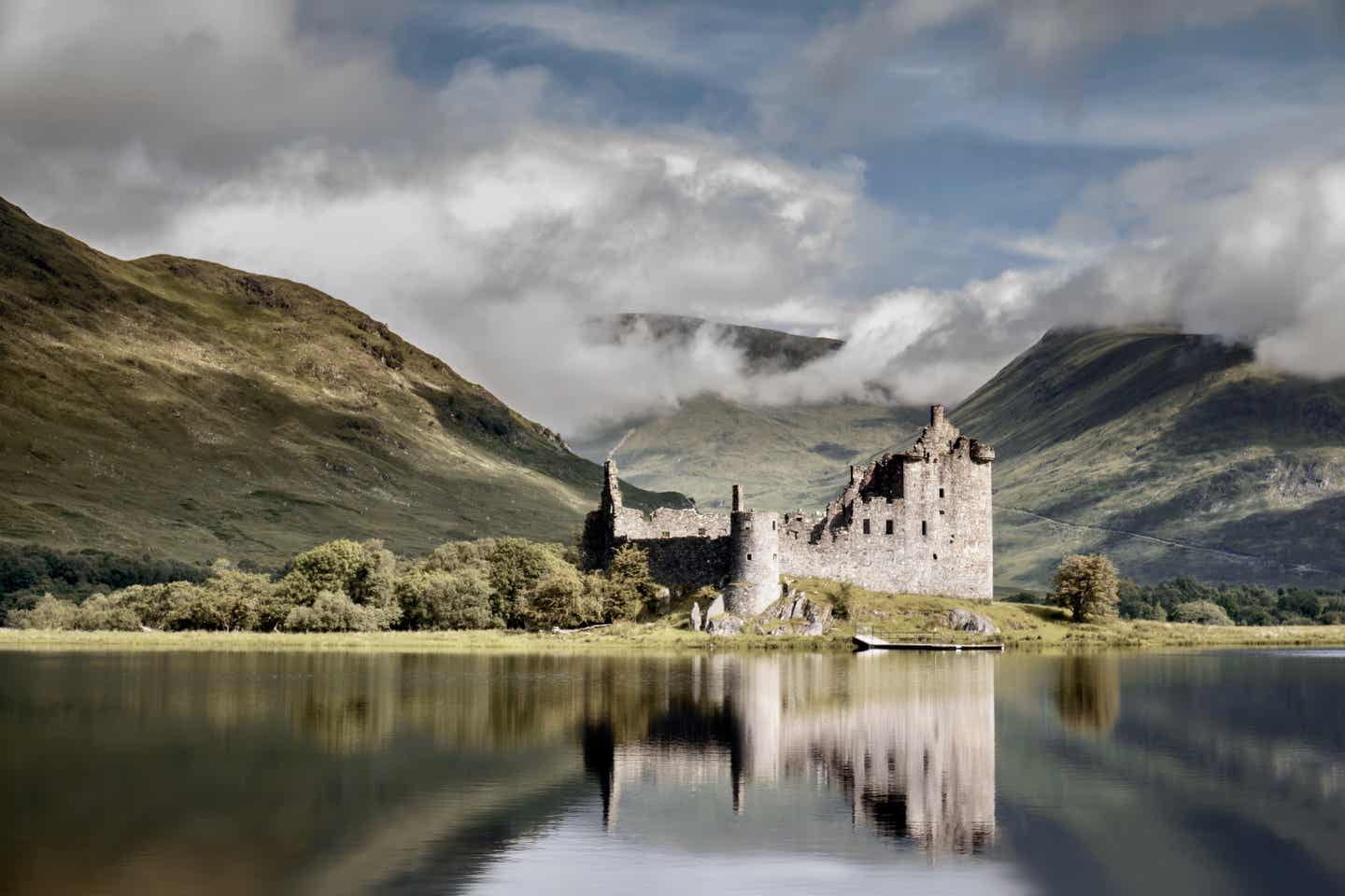 Blick auf das Kilchurn Castle in Schottland, das sich im Wasser des Loch Awe spiegelt; im Hintergrund wolkenverhangene Highlands