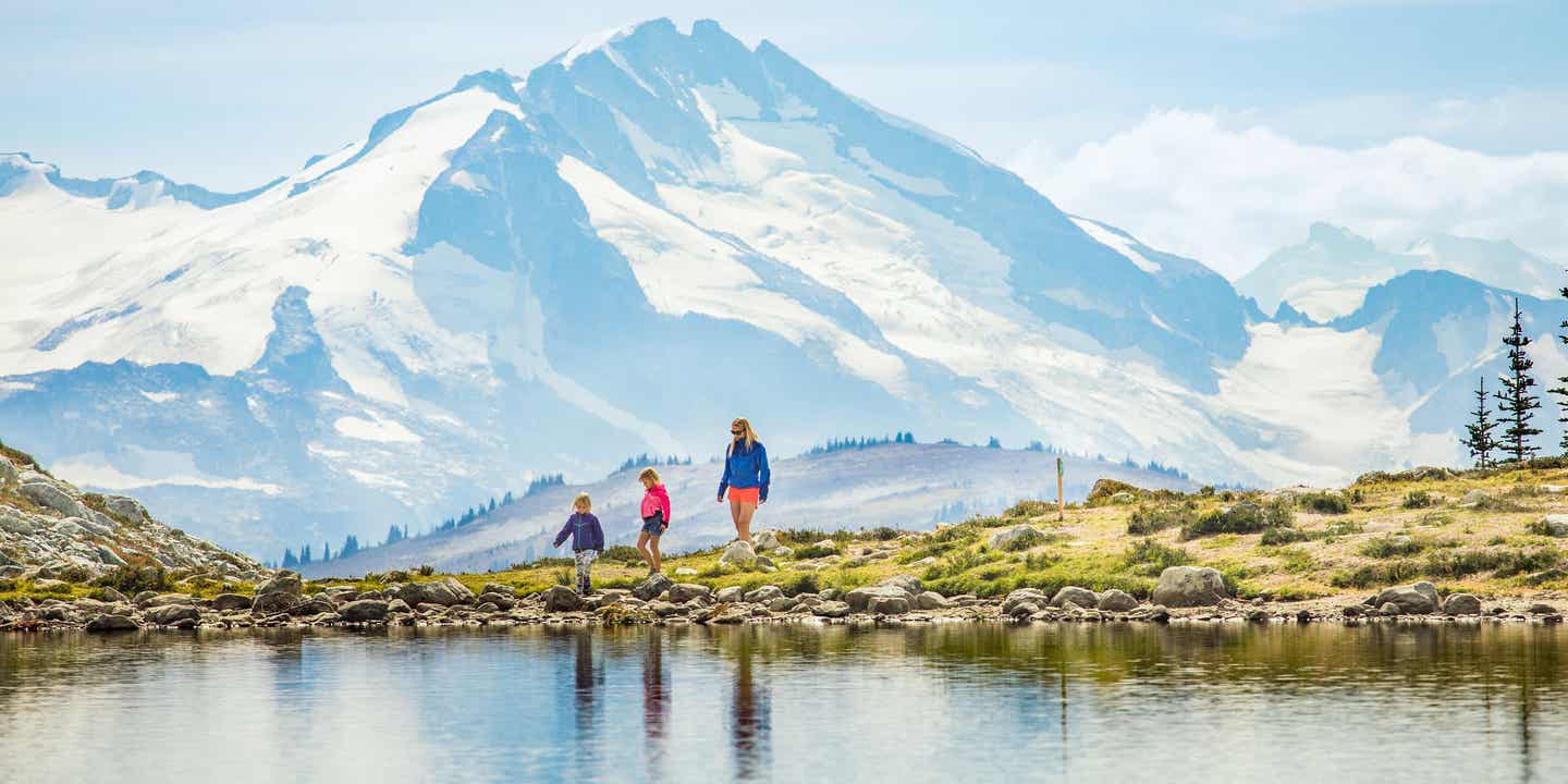 Eine Mutter wandert mit zwei Kindern an einem See entlang, im Hintergrund schneebedeckte Berge