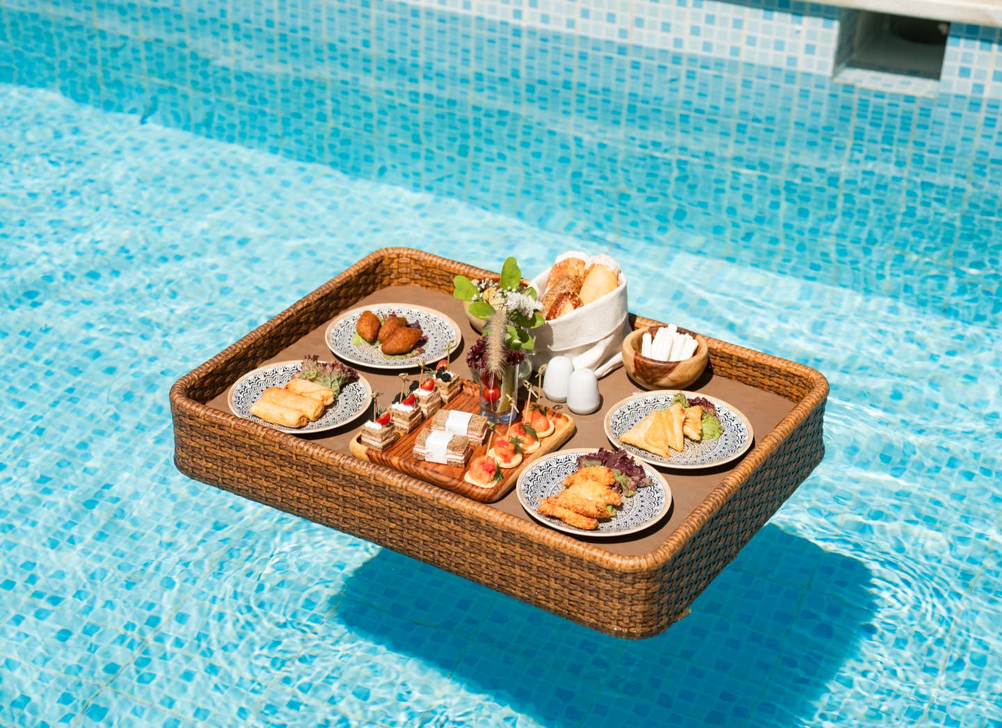Geflochtener Tablettkorb mit verschiedenen Frühstücksgerichten schwimmend im Pool