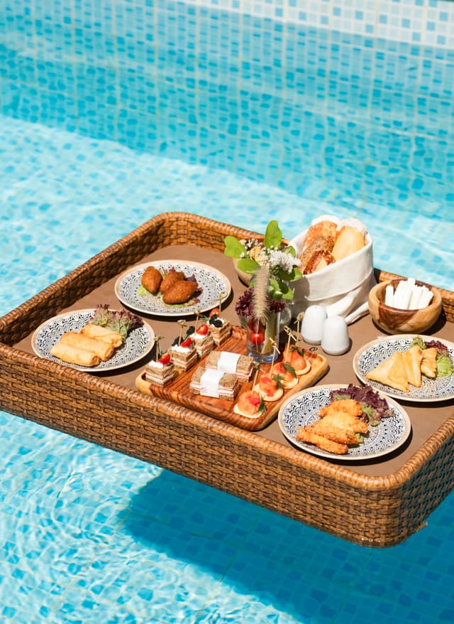 Sentido Gastronomie Geflochtener Tablettkorb mit verschiedenen Frühstücksgerichten schwimmend im Pool
