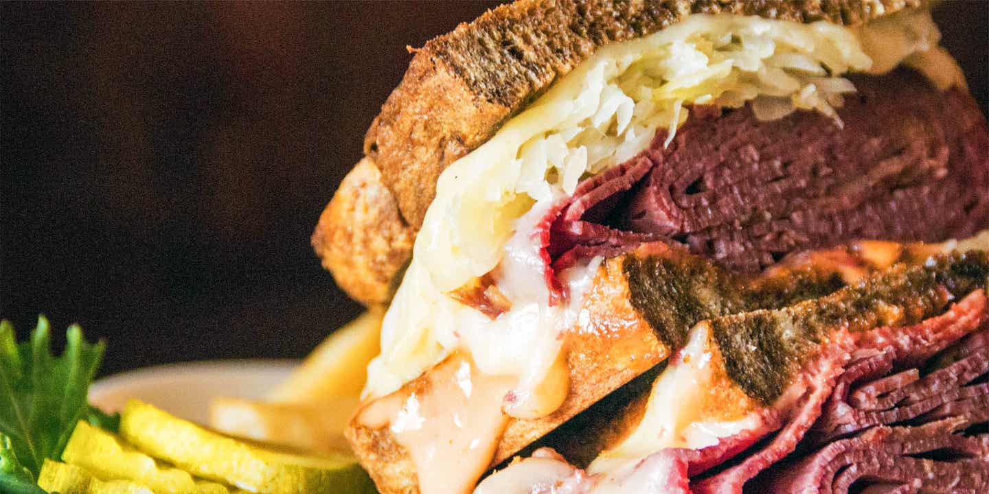 Ein üppig mit Pastrami und Sauerkraut belegtes Sandwich mit eingelegte Gurken daneben vor einem schwarzen Hintergrund.