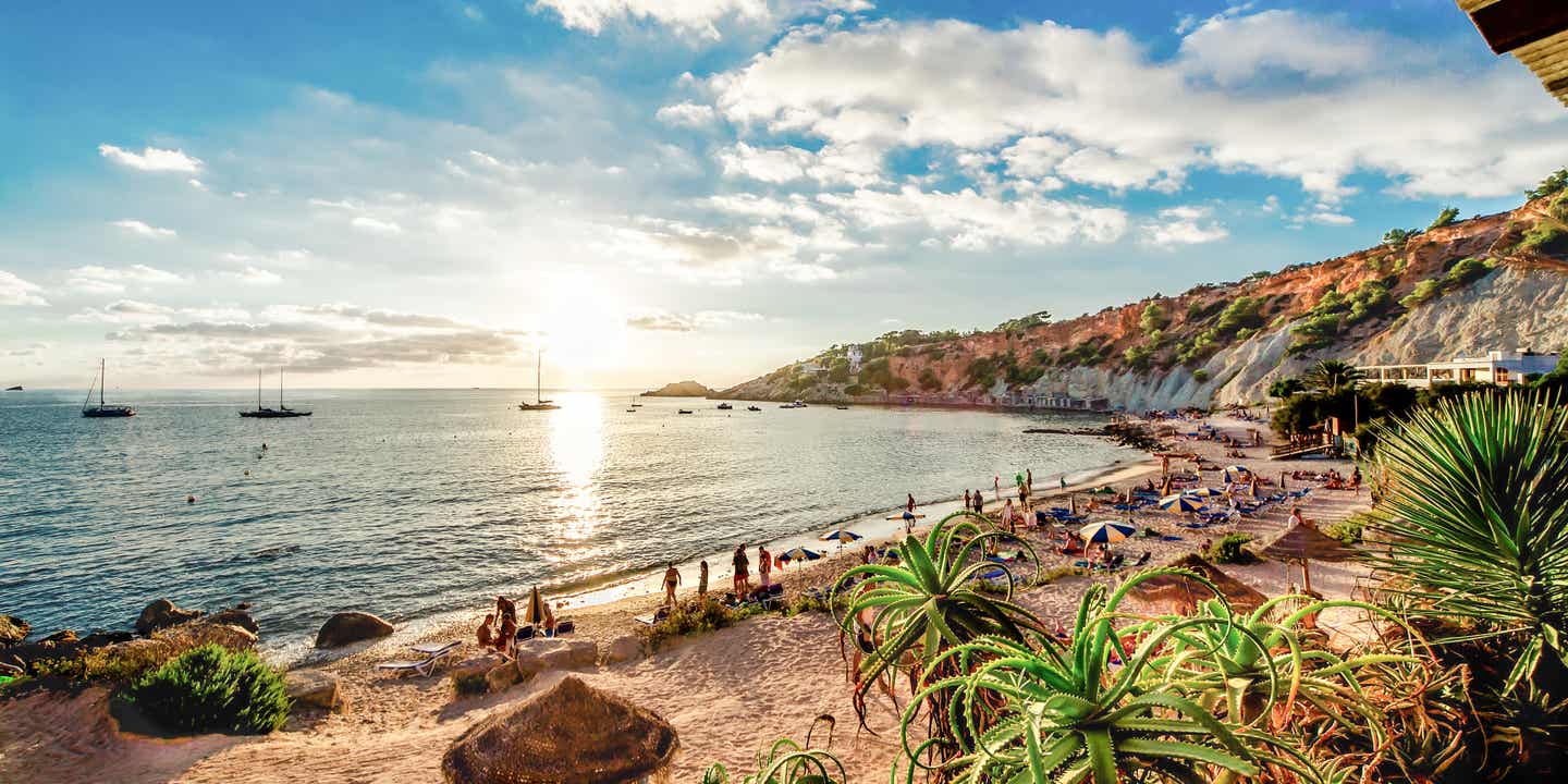 Sonnenuntergang über dem Strand von Cala d'Hort auf Ibiza mit Blick auf Liegen, Palmen und die Uferterrasse im Abendlicht 