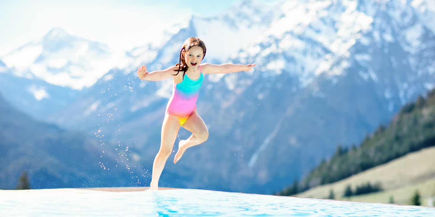 Ein Mädchen springt vom Rand eines Infinity-Hotelpools ins Wasser; im Hintergrund eine verschneite Bergkulisse