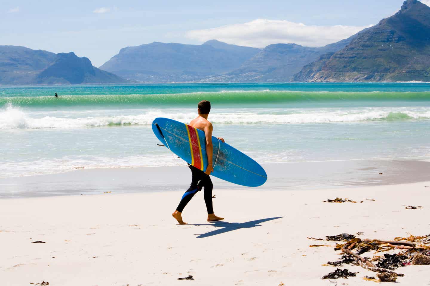 Beste Reisezeit Südafrika: Mann am Strand trägt Surfbrett