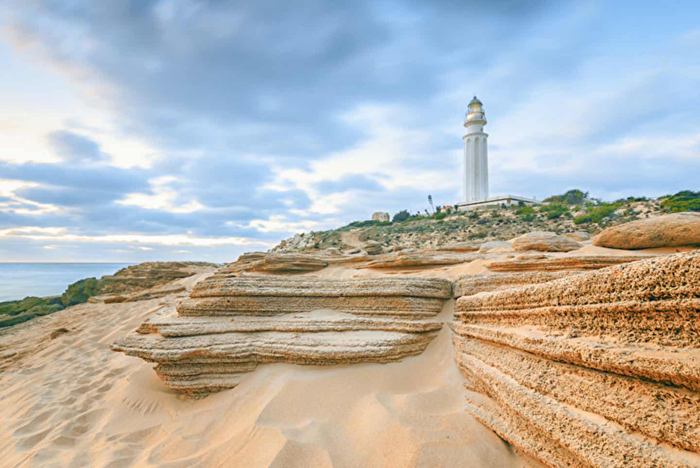 Sehenswürdigkeit Andalusiens: Leuchtturm am Kap Trafalgar bei Sonnenuntergang, umgeben von Sanddünen und Felsplatten