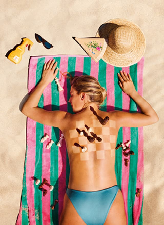Angebote entdecken Last Minute 2026: Frau liegt auf einem Strandtuch im Sand, auf ihrem Rücken ist ein Schachbrett mit Figuren. Neben ihr liegen Sonnencreme, Hut, Brille und Fächer