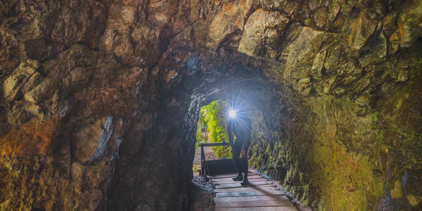 Wanderer mit Lichtquelle in engem Levada-Tunnel auf Madeira, feuchte Felswände und schmaler Steg