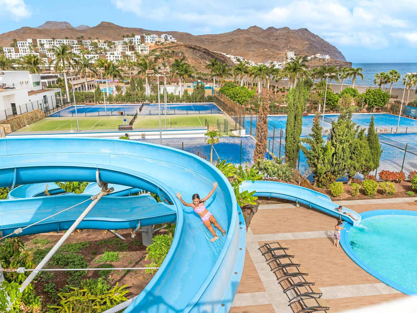 Kind rutscht auf blauer Wasserrutsche im Playitas Aparthotel mit Blick auf Tennisplätze und das Meer