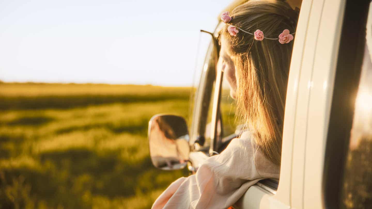 Frau mit einem Blumenkranz im Haar blickt aus dem Auto auf den Sonnenuntergang