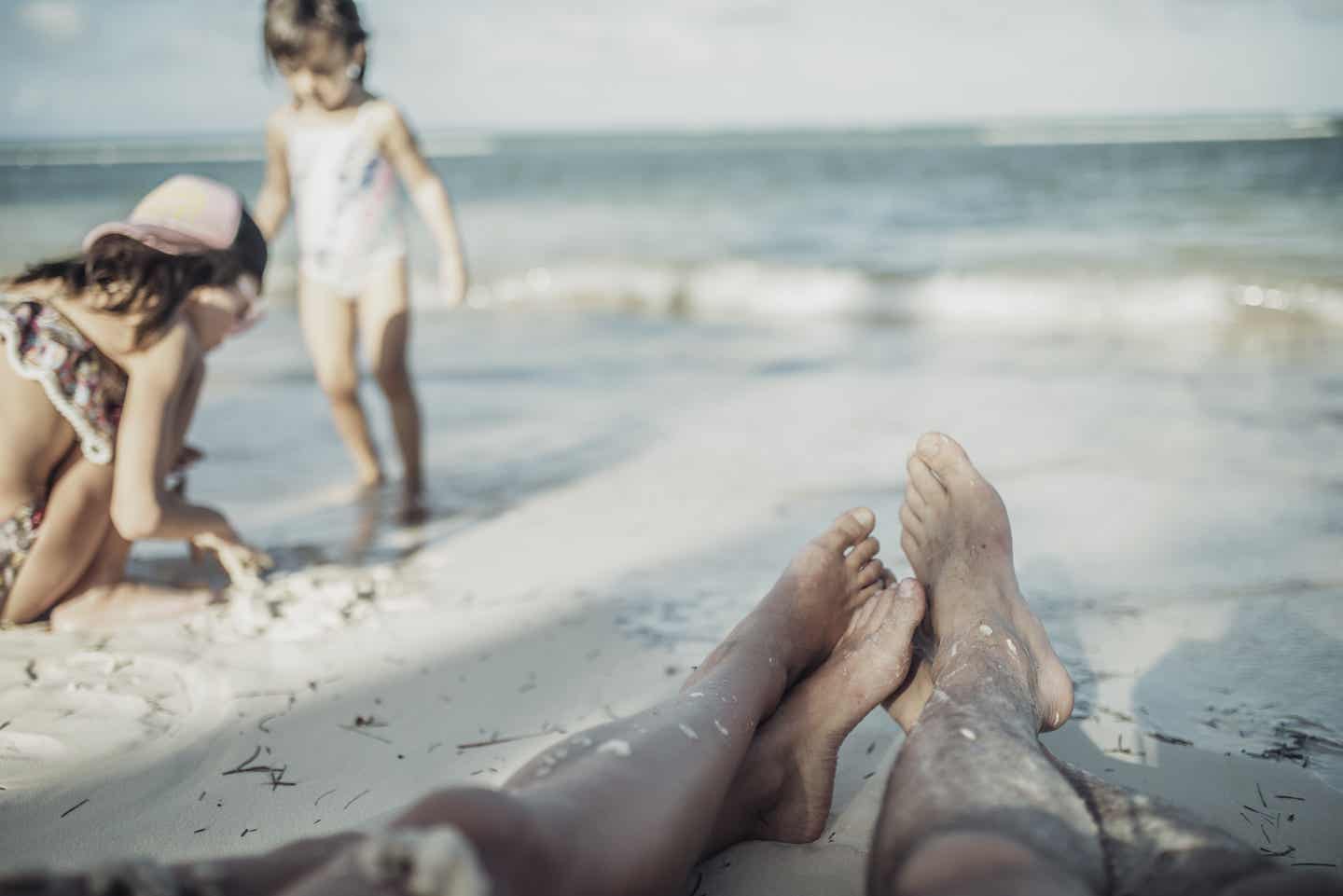 Puerto Plata Urlaub mit DERTOUR. Paar streckt am Karibik-Strand die Füße aus, während daneben die beiden Kinder im Sand spielen