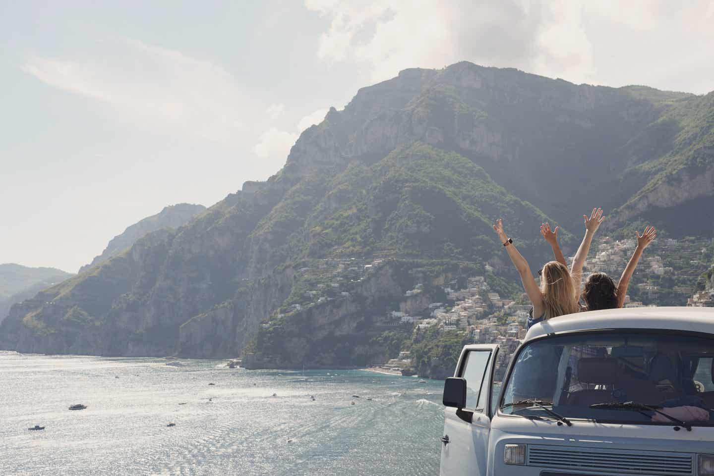 Kalabrien-Urlaub: Zwei Frauen vor einer Küstenlandschaft strecken ihre Arme aus einem Bulli heraus Richtung Himmel