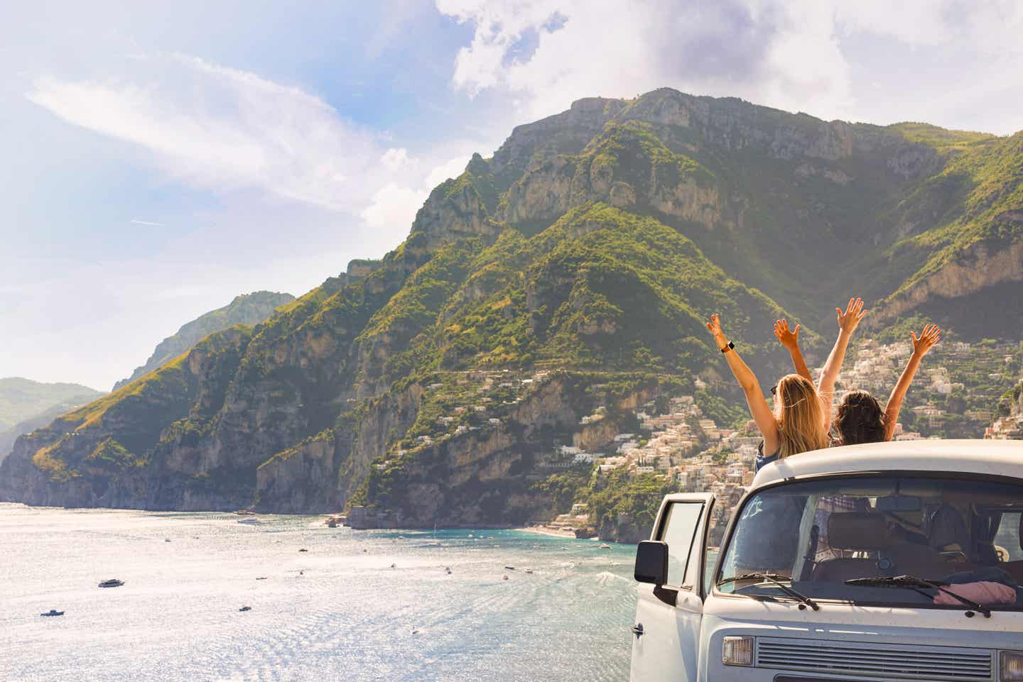 Kalabrien-Urlaub: Zwei Frauen vor einer Küstenlandschaft strecken ihre Arme aus einem Bulli heraus Richtung Himmel