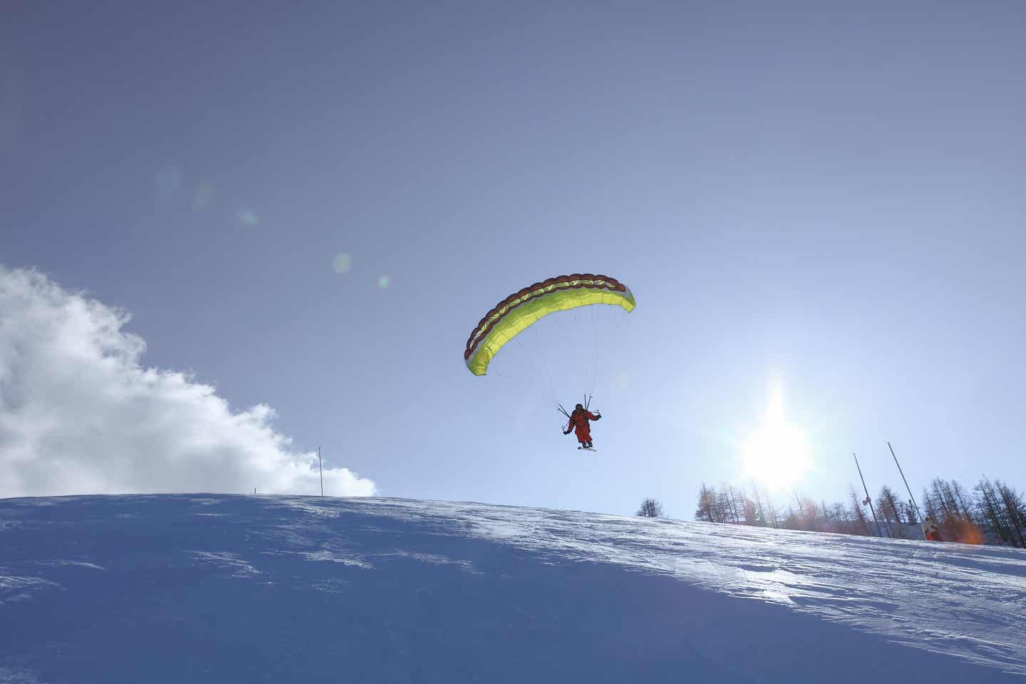 Mann mit Skiern und Schirm fliegt über einen schneebedeckten Hügel