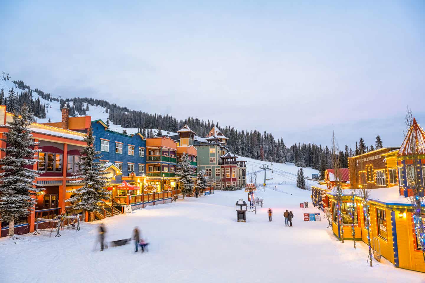 Abendstimmung in einem Skiresort, das direkt an einem Skilift gelegen ist, mit beleuchteten Häusern und Alpinisten auf der Straße