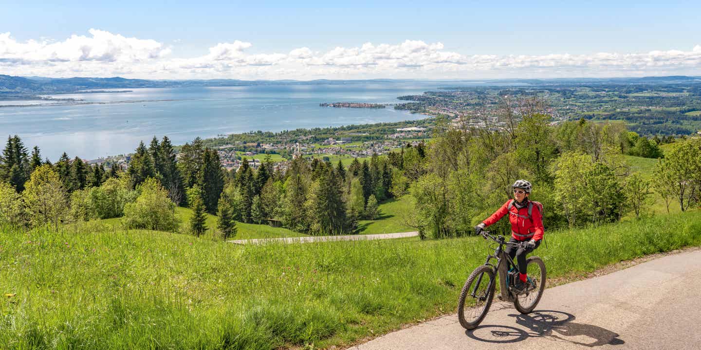 Frau radelt oberhalb des Bodensees mit Blick auf die Alpen unter blauem Himmel