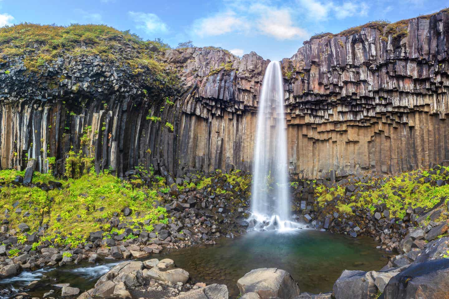 Zwischen kantig-geometrisch geformten dunklen Lavafelsen stürzt sich das Wasser des Svartifoss auf Island in die Tiefe.