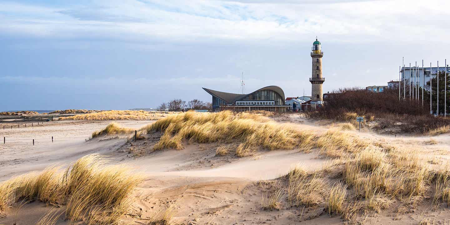 Leuchtturm und Teepott in Warnemünde, umgeben von Dünenlandschaft und Ostseestrand, unter einem leicht bewölkten Himmel