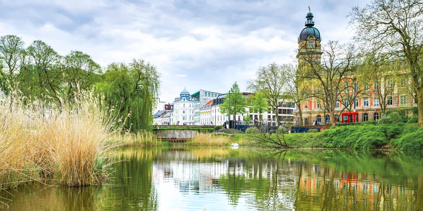 Grüne Uferlandschaft im Park Planten un Blomen mit Blick auf Wasser, Bäume und historische Gebäude in Hamburg