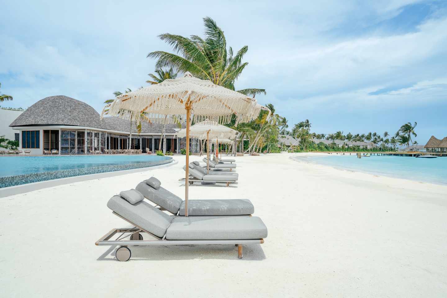 Strand mit Liegestühlen und Sonnenschirmen vor einem Pool und tropischen Bäumen unter bewölktem Himmel