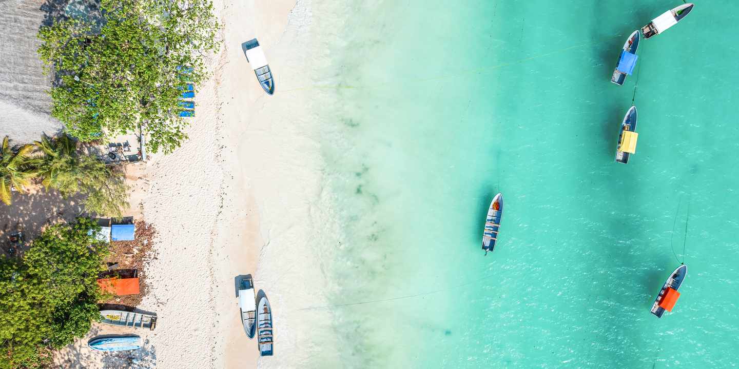 Luftaufnahme von Kizimkazi mit hellem Sandstrand, grünen Bäumen und farbigen Fischerbooten, die im klaren Wasser treiben 