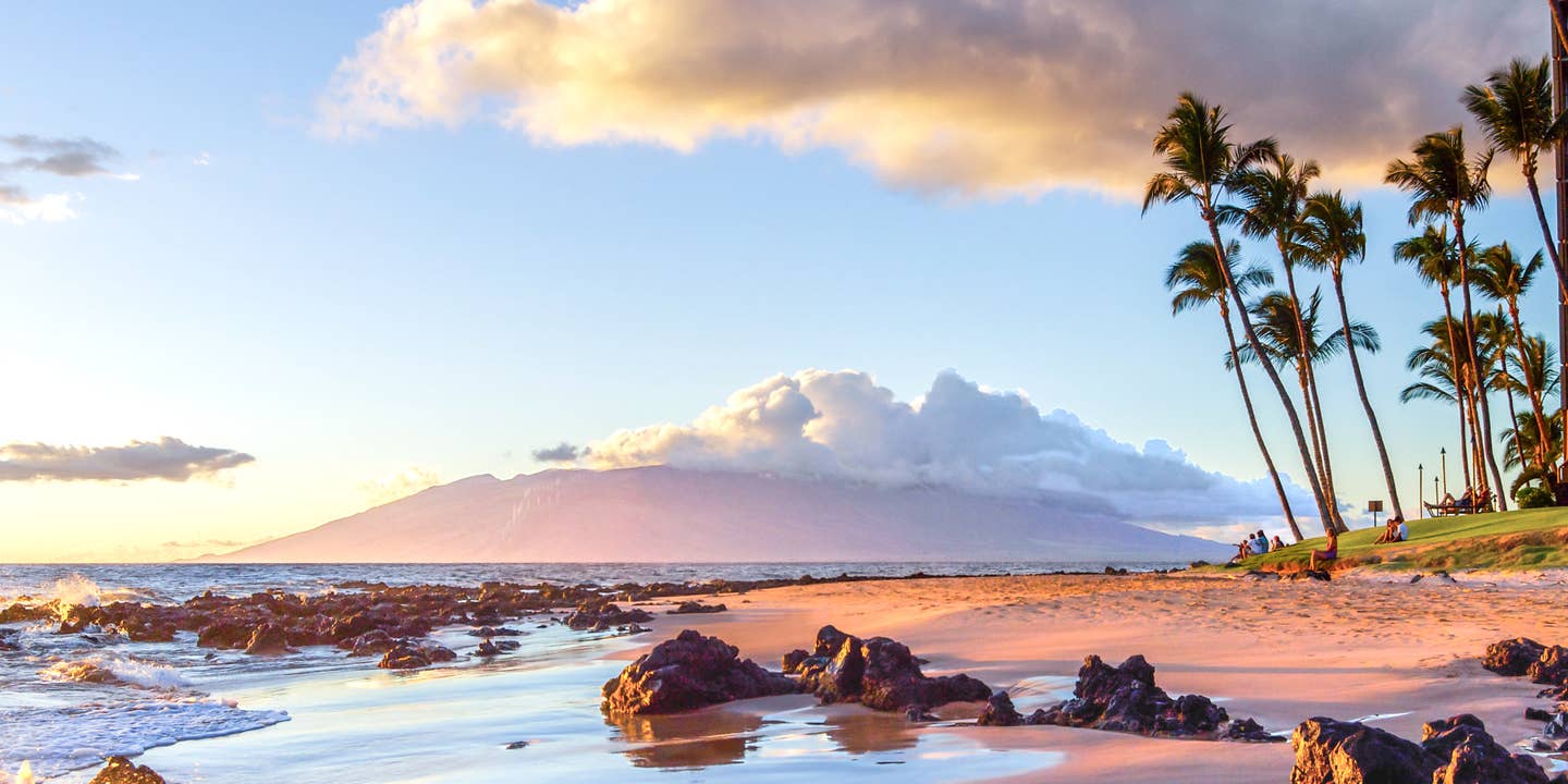 Sonnenunterganz am Strand von Maui im Hawaii-Urlaub; im Hintergrund ein wolkenverhangener Berg auf einer Nachbarinsel.
