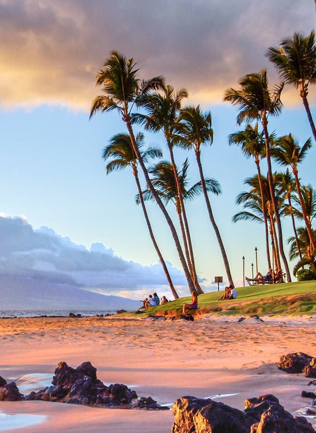 Hawaii Urlaub Sonnenunterganz am Strand von Maui im Hawaii-Urlaub; im Hintergrund ein wolkenverhangener Berg auf einer Nachbarinsel.