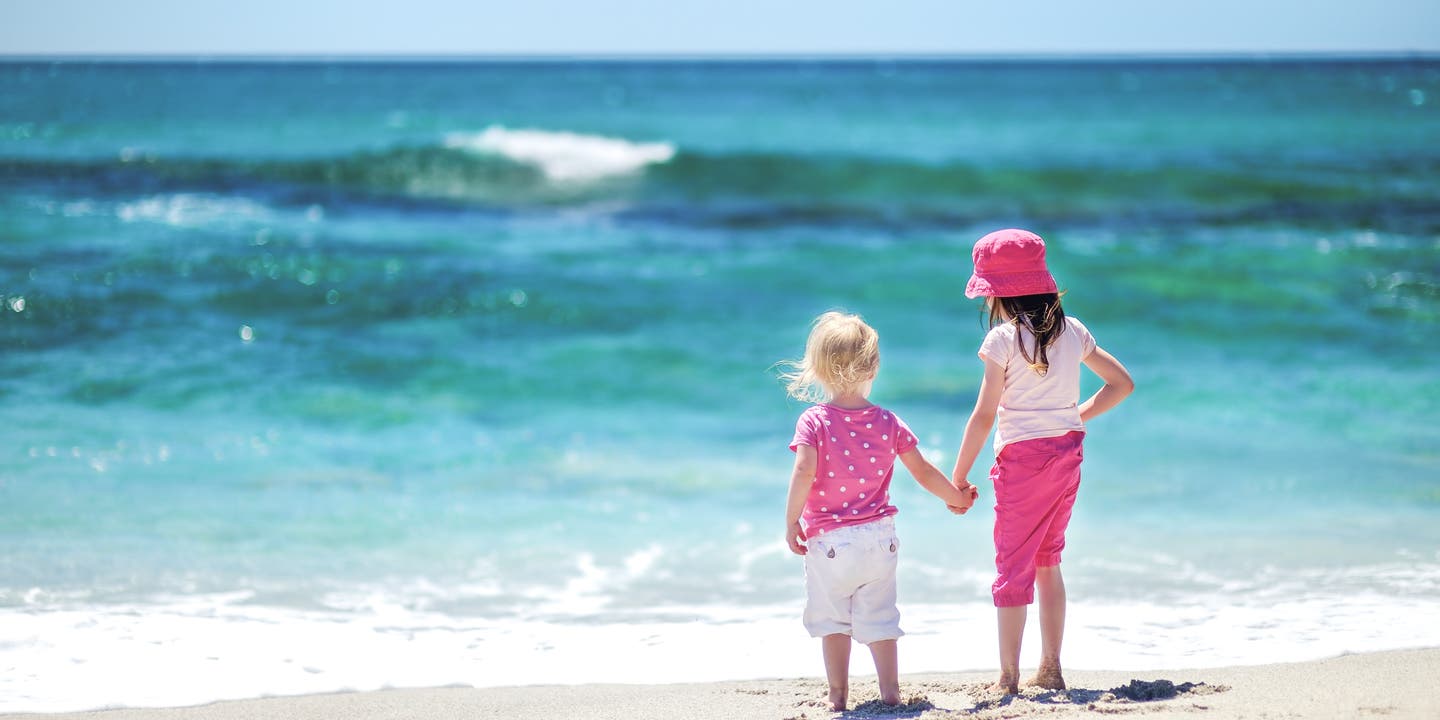 Zwei Mädchen stehen mit dem Rücken zur Kamera am Strand, im Hintergrund ist das Meer.